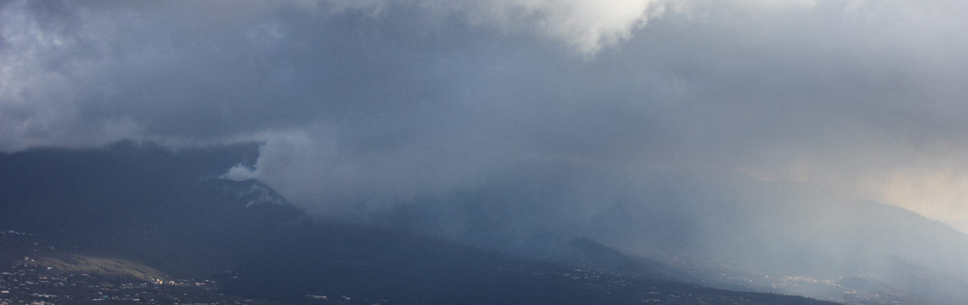 Volcanic mountain shrouded in mist and clouds, with agricultural fields and scattered settlements visible in the valley below under dramatic sky.