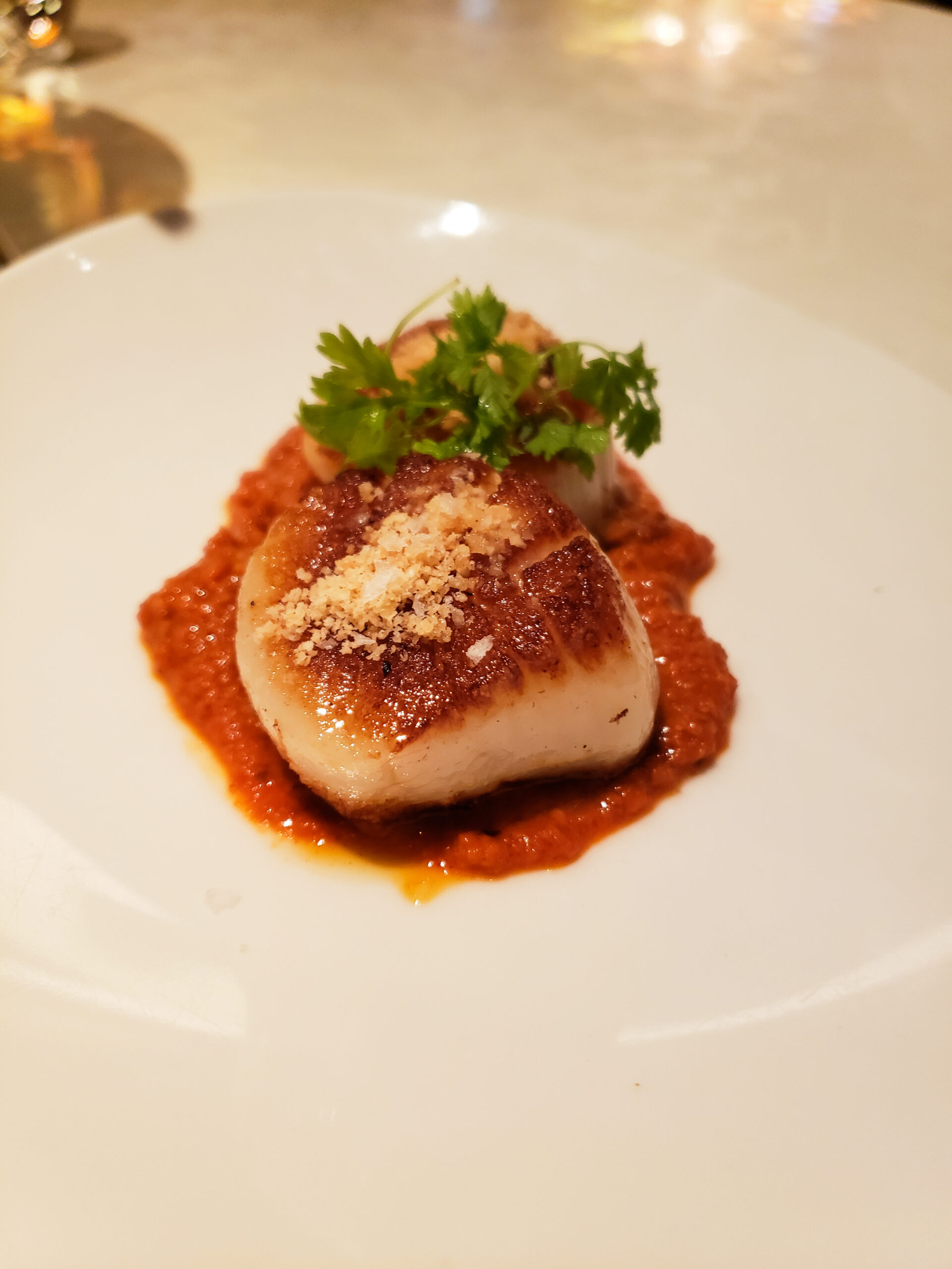 A perfectly seared scallop topped with crispy breadcrumbs and fresh cilantro, served on a vibrant red mojo sauce.