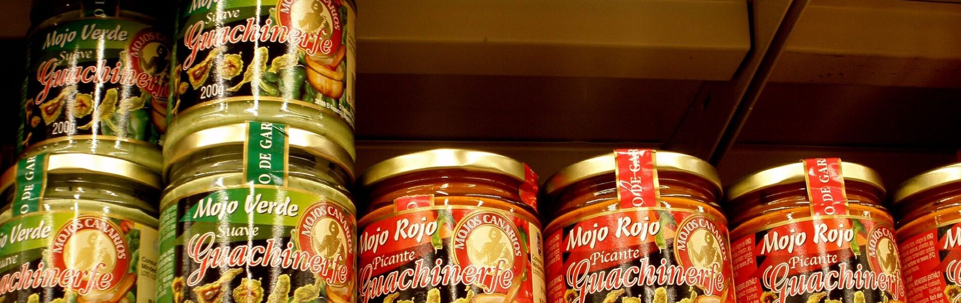 Glass jars of mojo rojo and mojo verde sauces with gold lids and colorful labels arranged on a shelf.