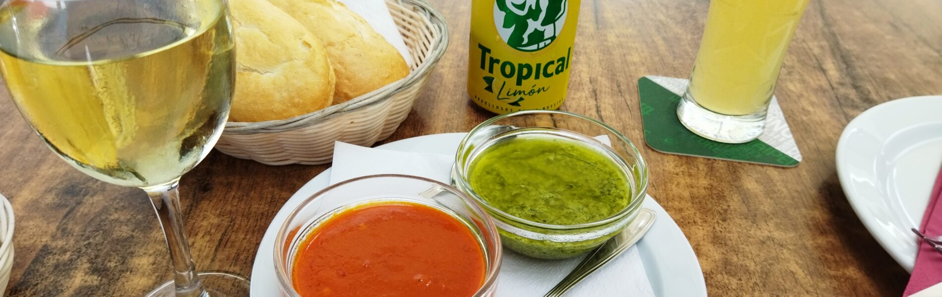 Outdoor table setting with two small bowls of sauce—red mojo and green mojo—beside bread basket, wine glass, tropical lemon beverage, and yellow drink
