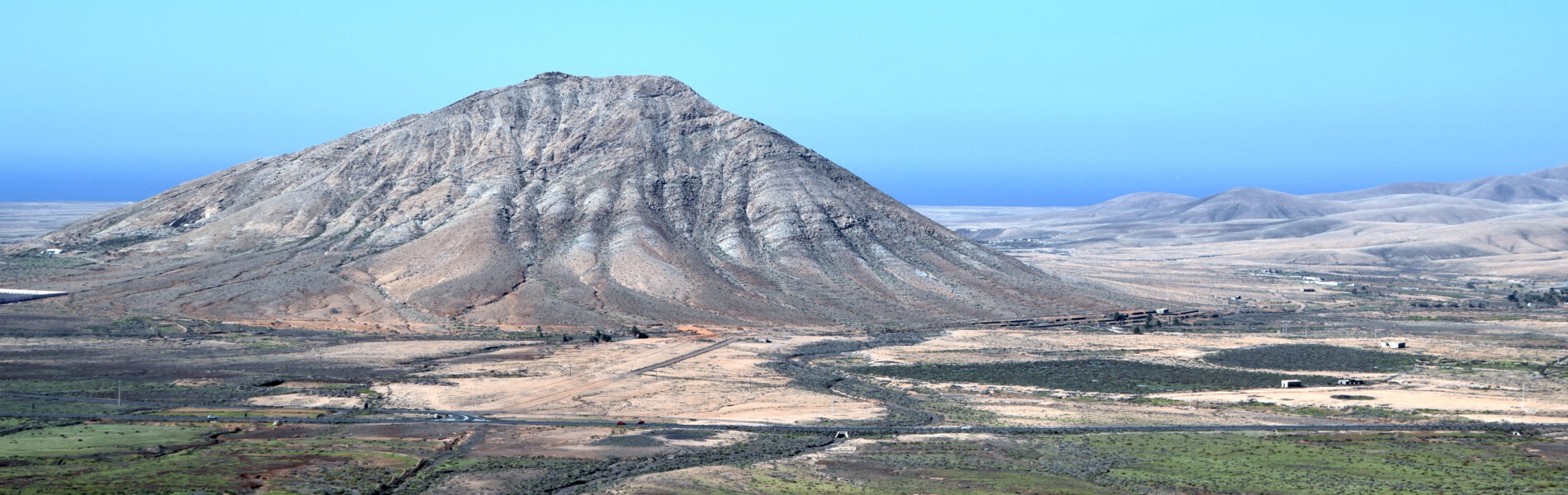 Montaña de Tindaya, a symmetrical volcanic cone with deep erosion gullies, rises above surrounding green agricultural plains and dark lava fields under clear blue sky.
