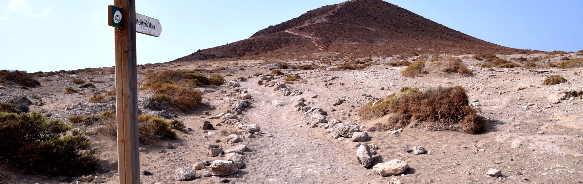 Wooden trail marker post on volcanic terrain with dark cinder cone ahead and sparse desert vegetation under clear blue sky