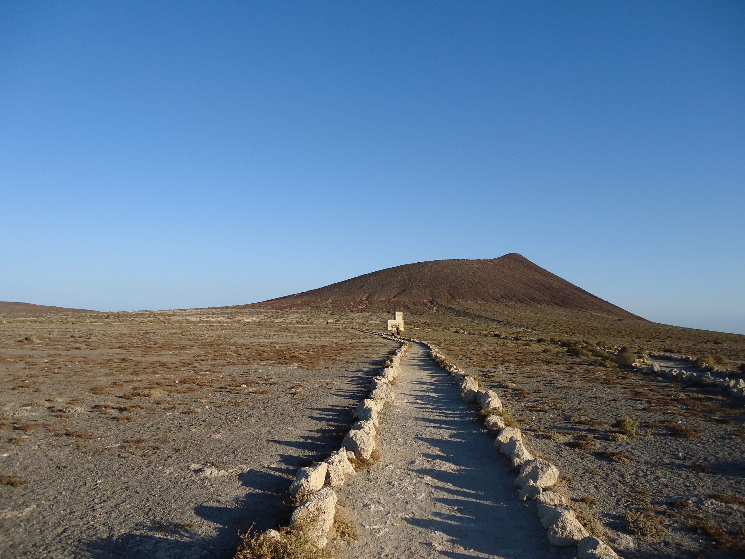 A stone-lined path crosses volcanic terrain toward a distinctive cinder cone under clear blue sky