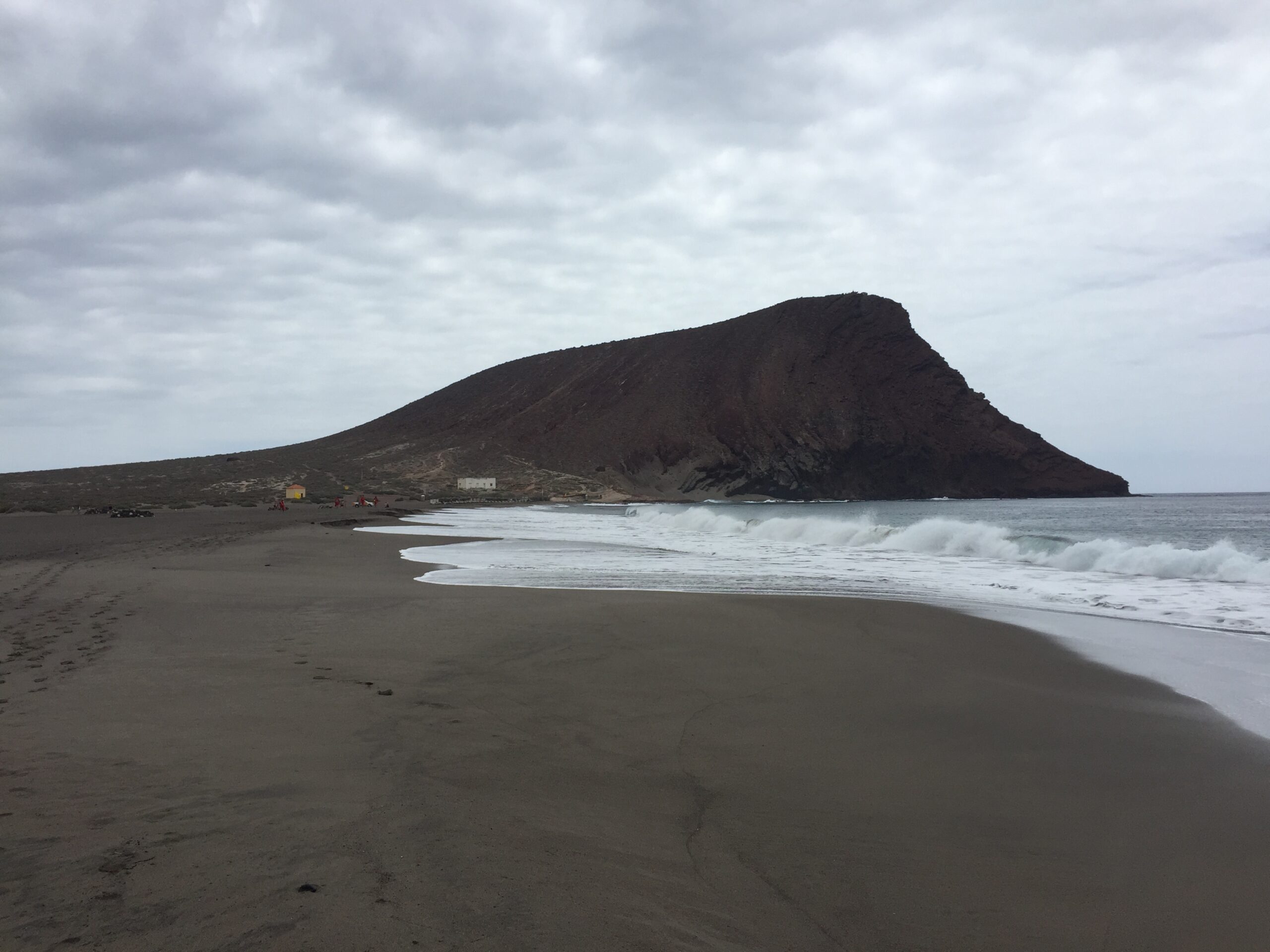 Dark volcanic cone rising above a black sand beach with white-capped waves and scattered buildings at its base under cloudy sky