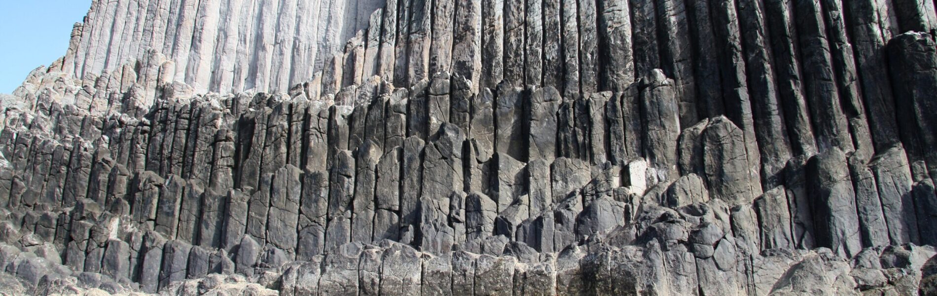 Massive hexagonal basalt columns rising from the sea, with weathered dark stone pillars stacked in geometric formations against a clear blue sky.