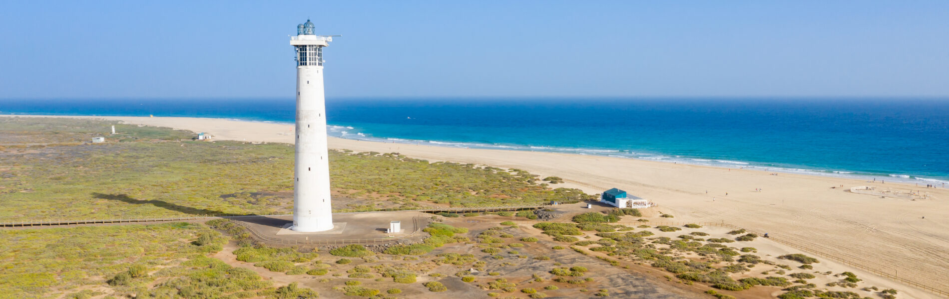 White cylindrical lighthouse on sparse, vegetation-covered terrain overlooking a wide sandy beach and turquoise ocean under clear blue sky.