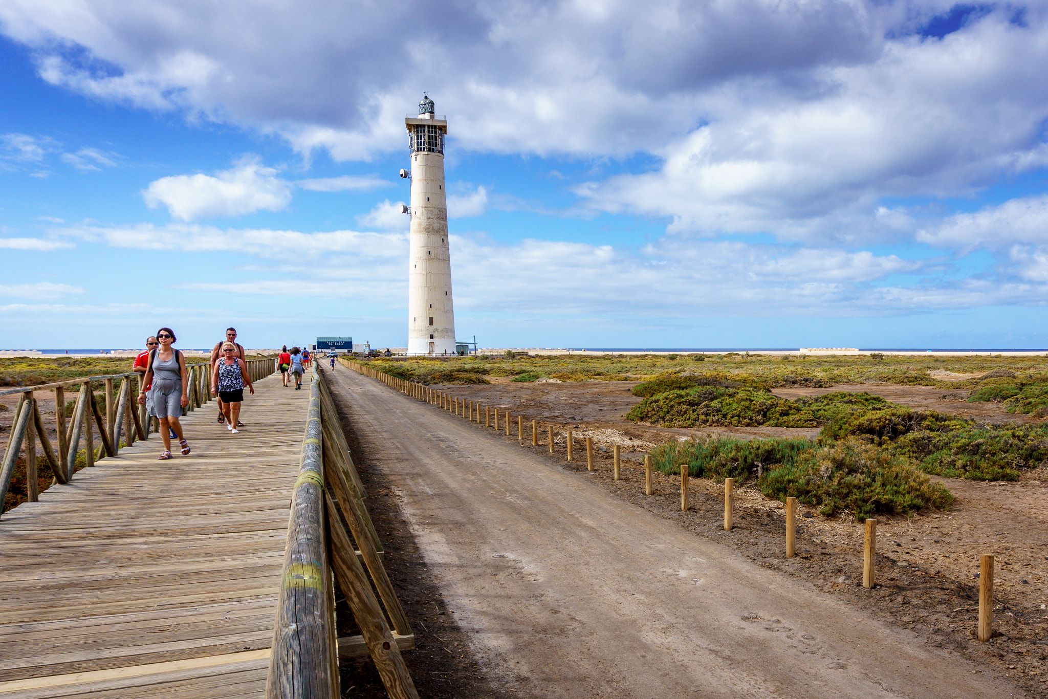 The distinctive white lighthouse at Morro Jable stands prominently across the arid landscape, accessible via a wooden boardwalk and sandy path.