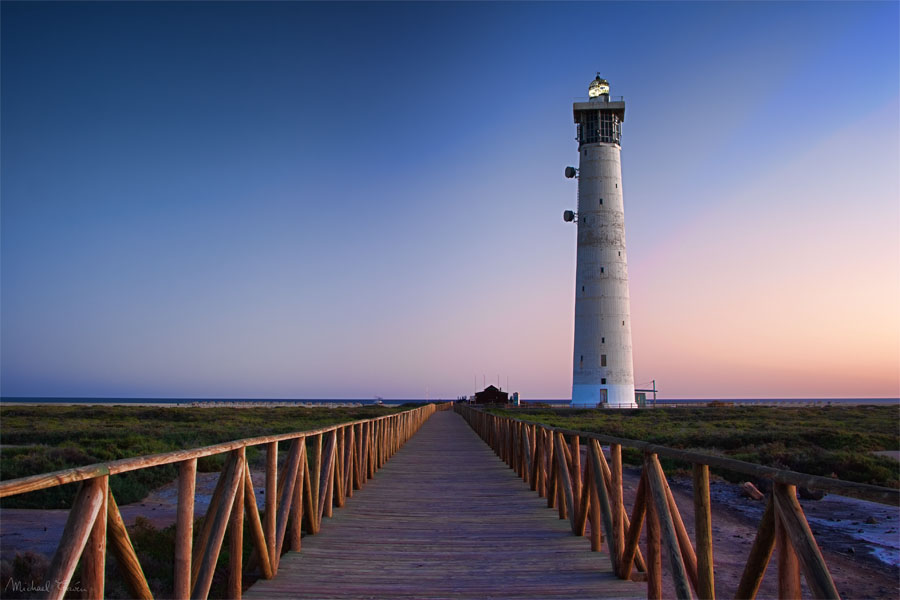 The Morro Jable Lighthouse stands at the southern tip of Fuerteventura, accessed by a wooden boardwalk that crosses the surrounding flat, arid landscape.