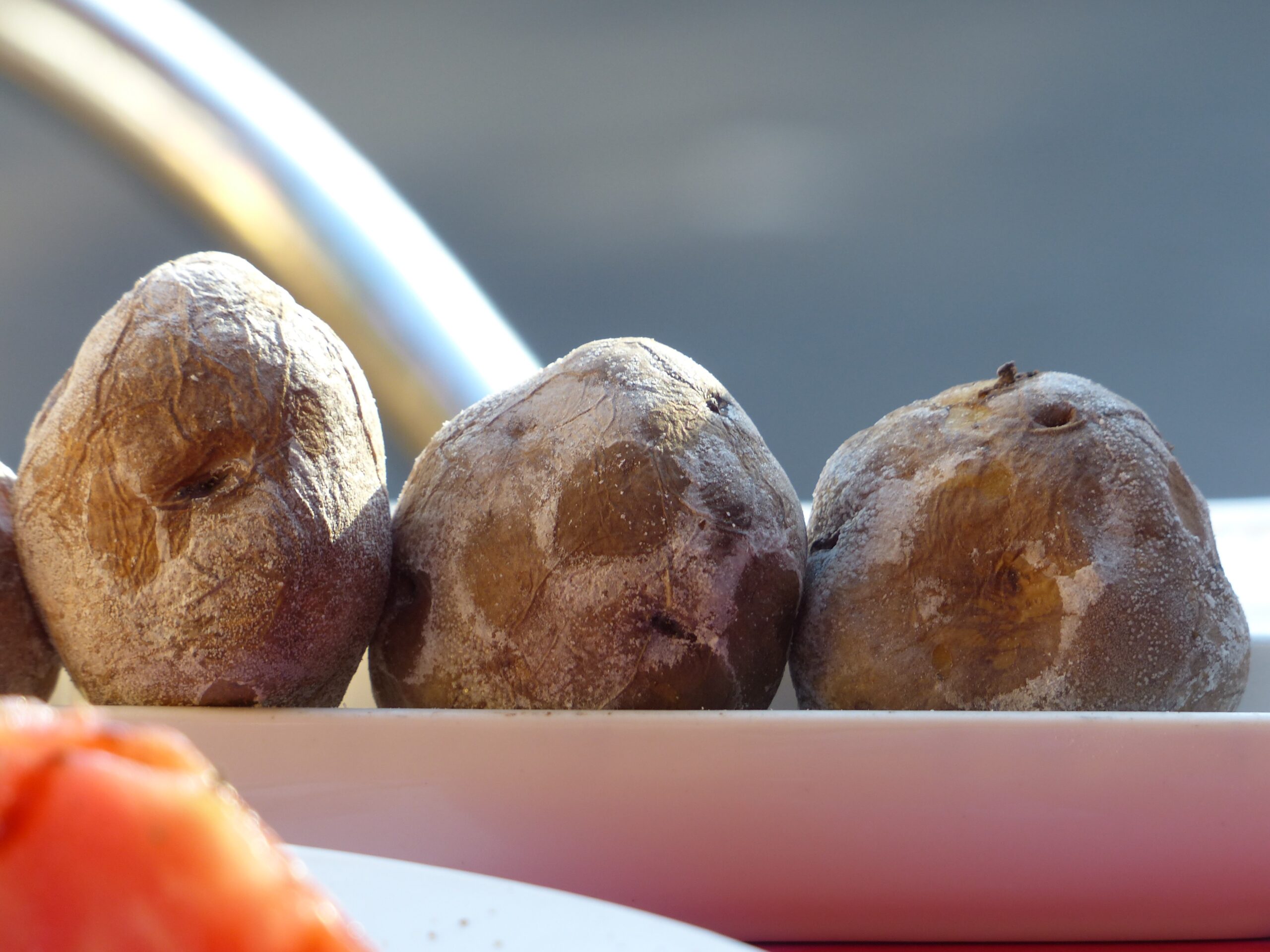 Three wrinkled potatoes with salt crust on a pink plate, bright sunlight casting shadows, blurred background.