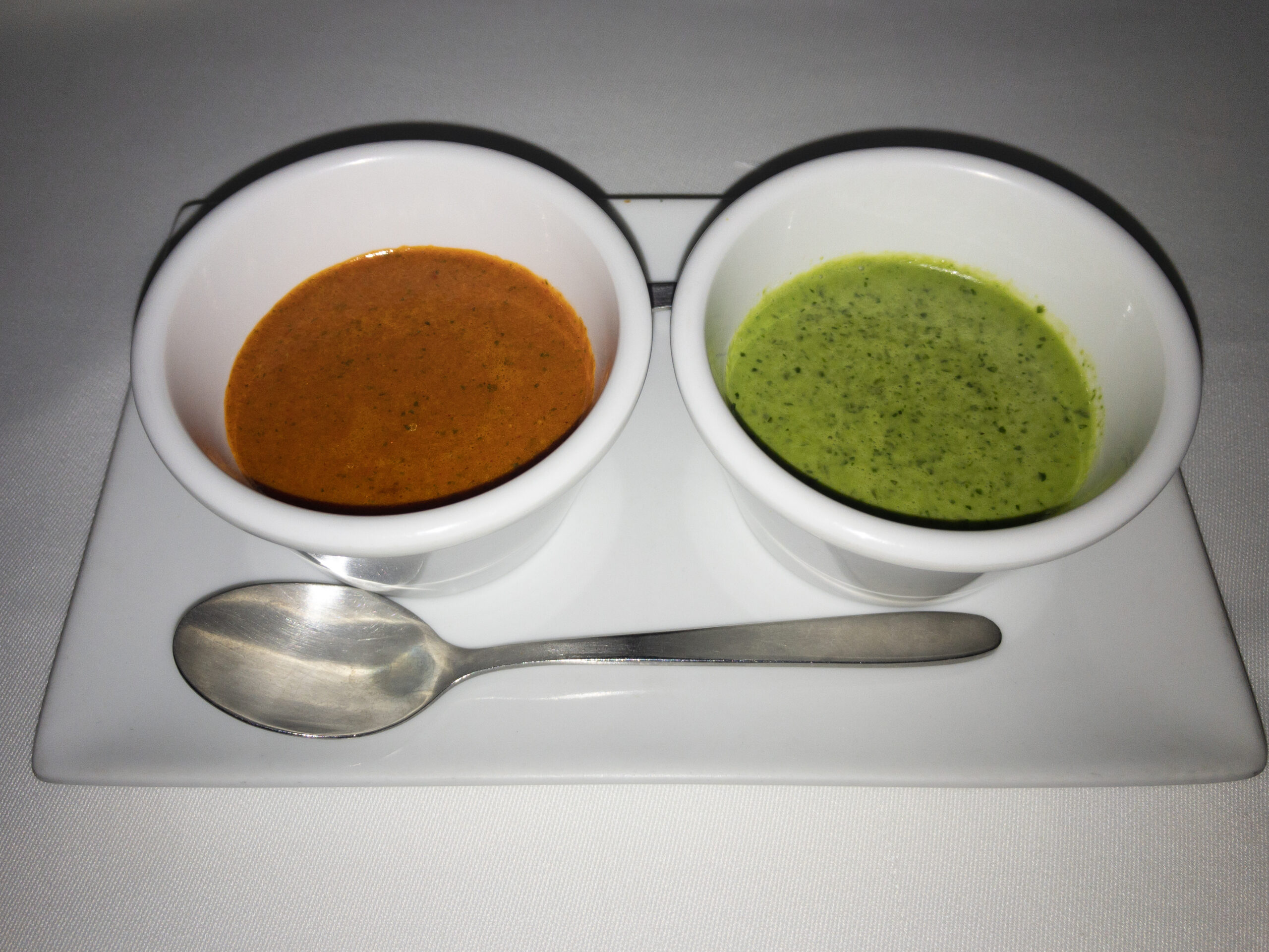 Two white bowls with brown and green sauces on a white rectangular plate with a metal spoon
