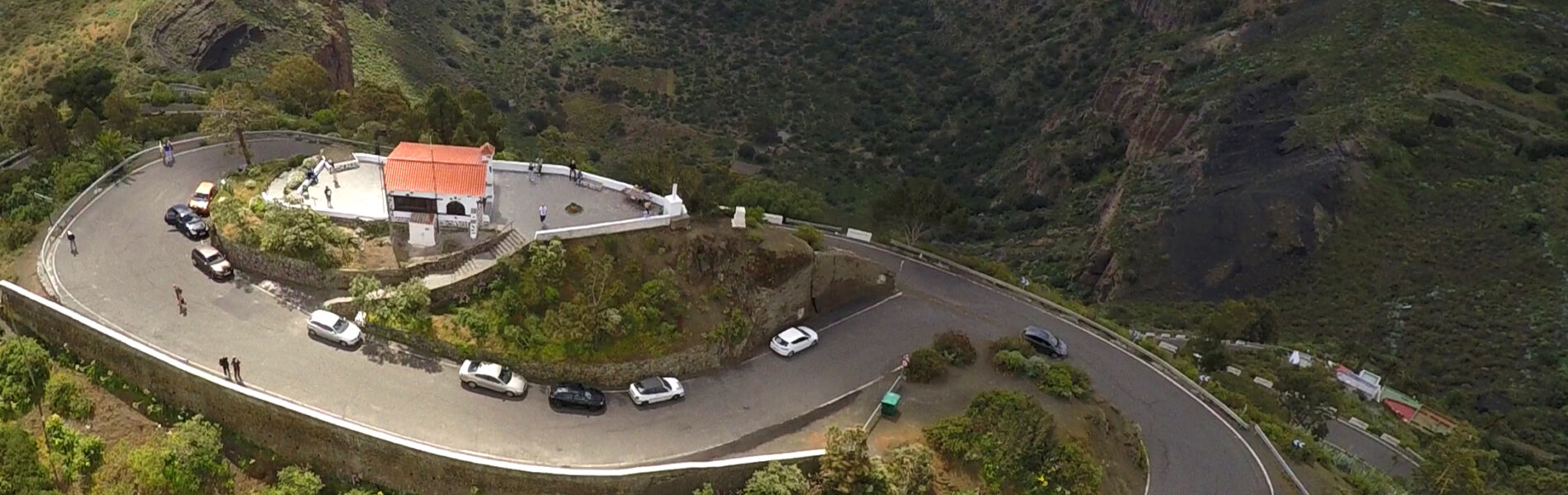 Aerial view of a hilltop viewpoint with white building and orange roof surrounded by a curved parking area, overlooking a deep volcanic crater with forested slopes.