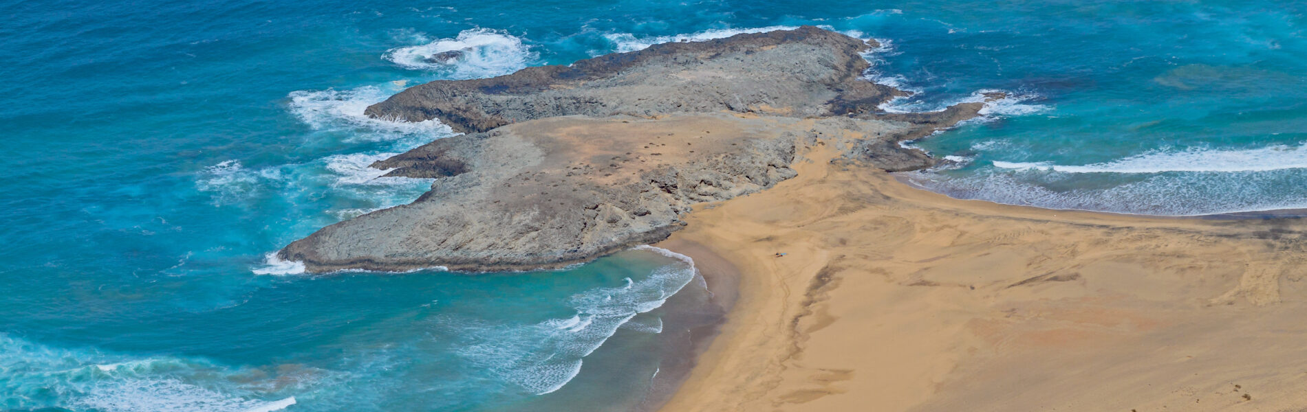 Golden sand beach with turquoise waves and dark rocky outcrops extending into the Atlantic Ocean under bright daylight