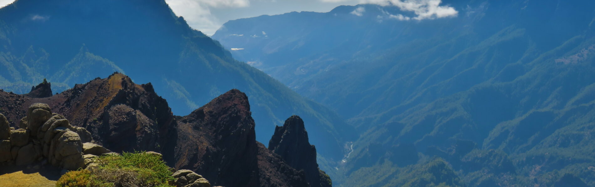 Rocky peaks and sparse vegetation in foreground overlook a deep valley with forested slopes and distant mountains under blue sky with clouds