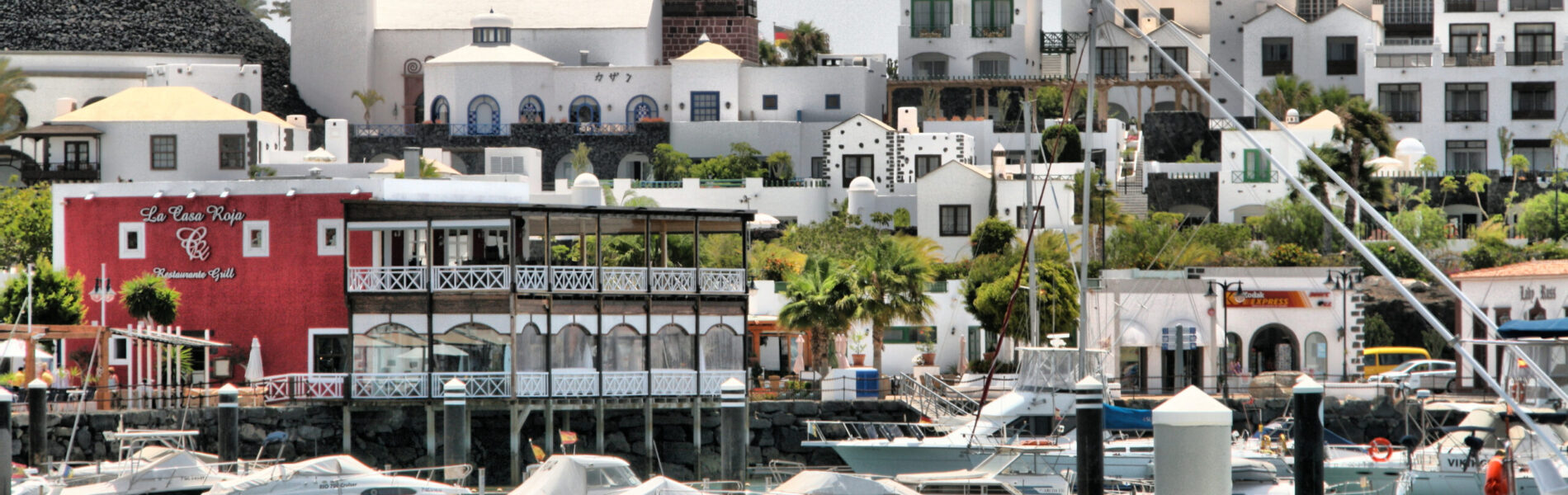 Waterfront harbor with moored boats, whitewashed buildings, a distinctive red restaurant, minaret tower, and palm trees under overcast sky
