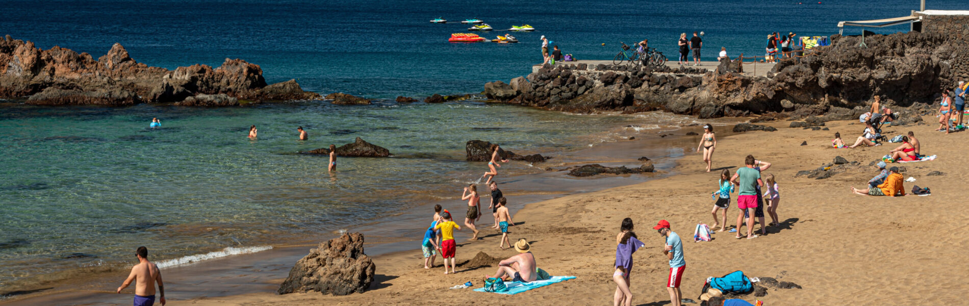 Sandy beach with swimmers and sunbathers, rocky outcrops, turquoise shallow water, and arid mountains across the blue sea under clear sky.