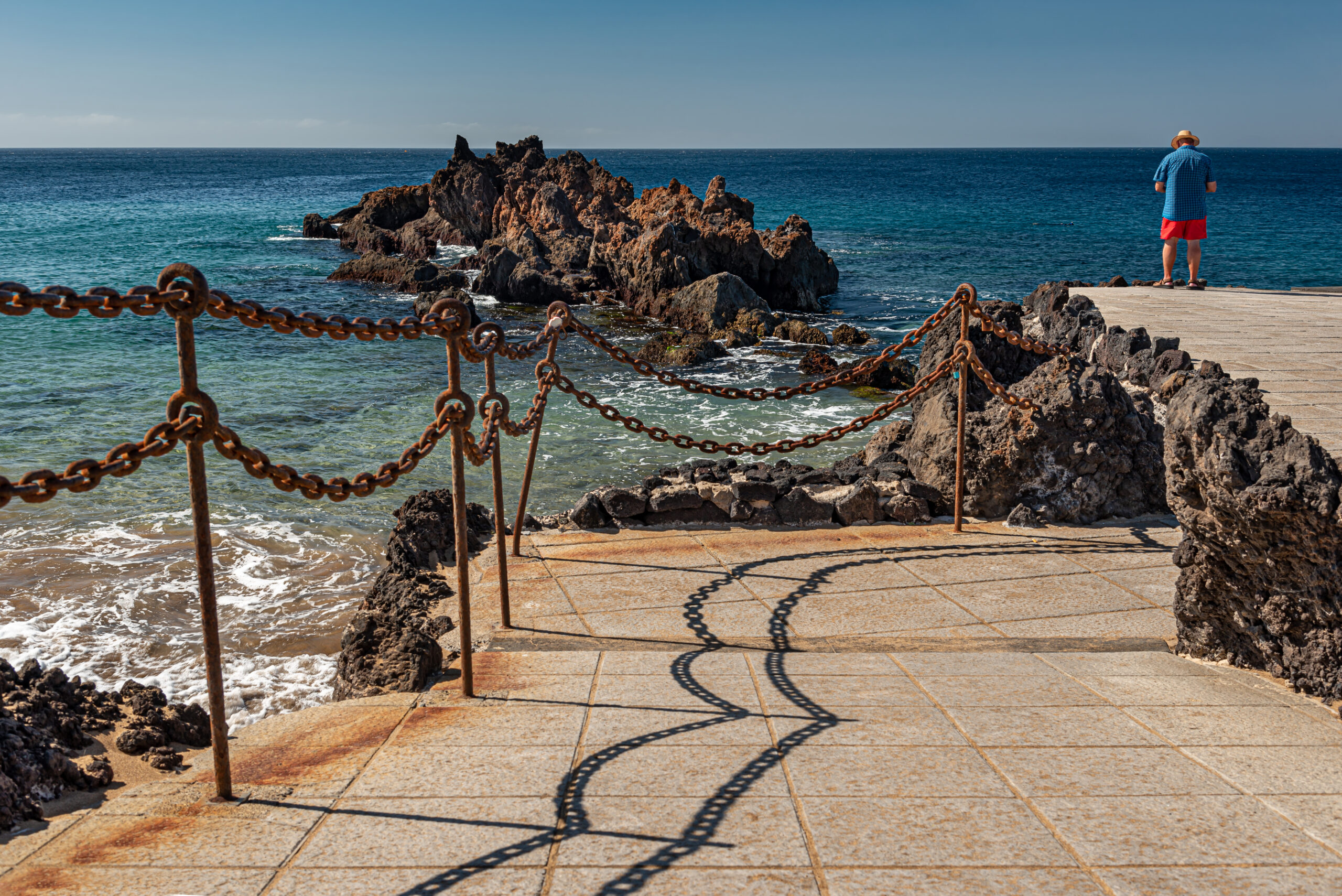A weathered chain barrier frames the scenic coastal walkway at Playa Chica, with dramatic rock formations visible in the clear Atlantic waters.