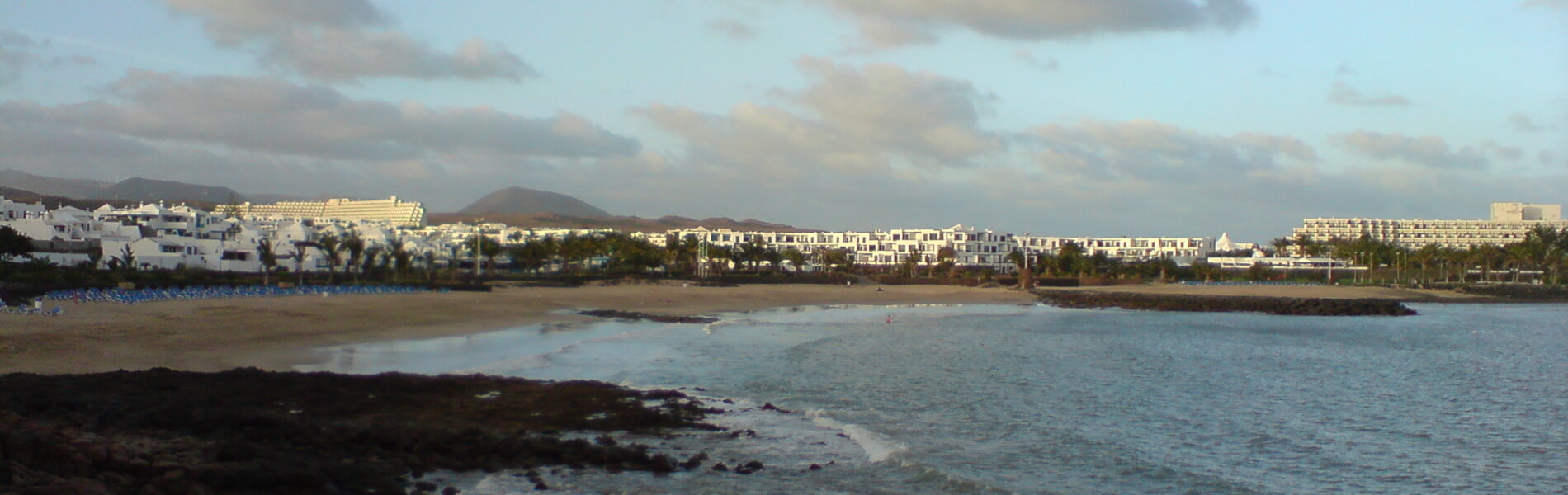 Rocky black volcanic shore with turquoise water and a sandy beach backed by white apartment buildings and a volcanic mountain under blue sky with scattered clouds.