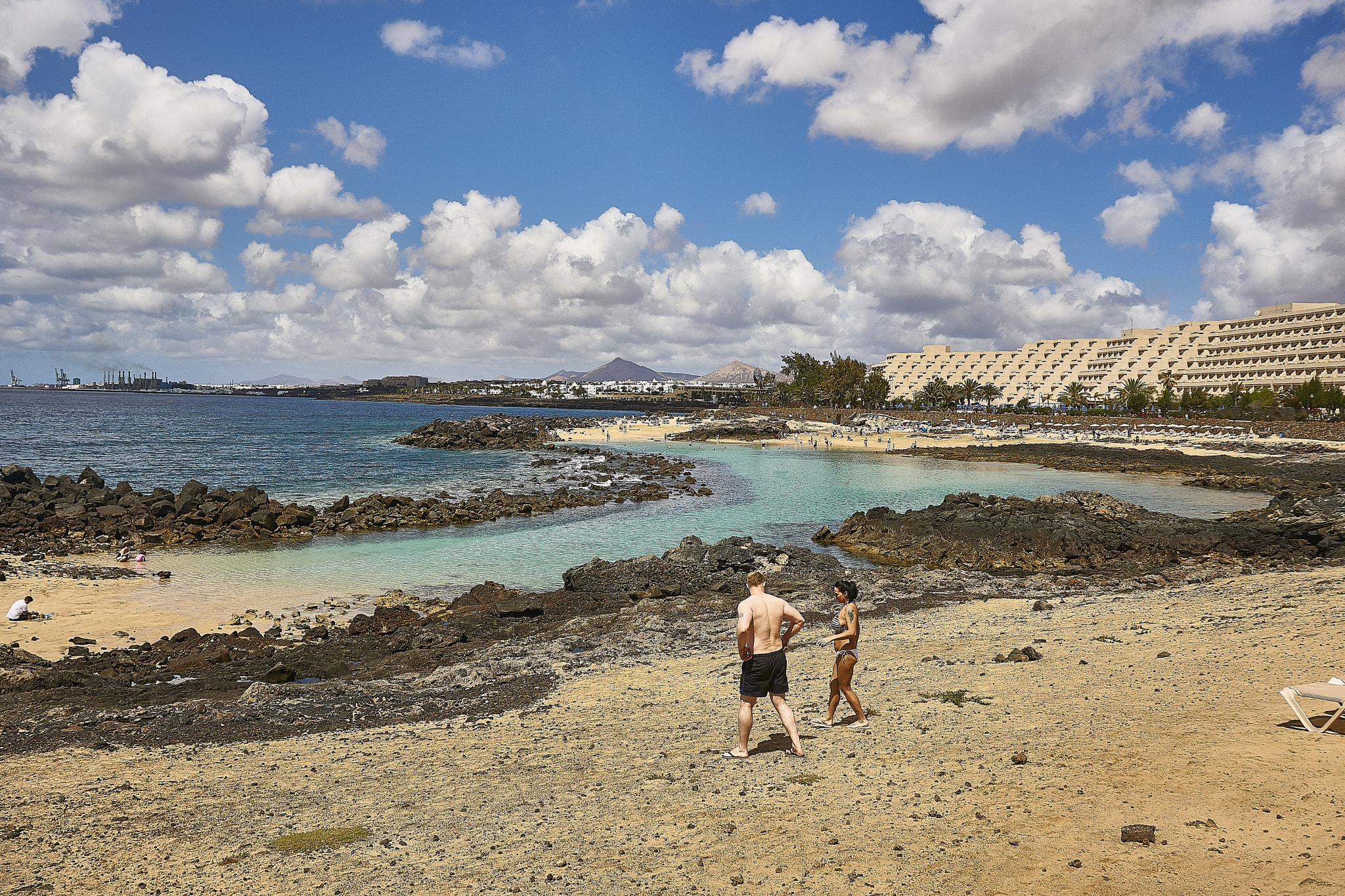 Playa de las Cucharas offers sheltered turquoise waters and dark volcanic rock formations, backed by the distinctive architecture of Costa Teguise's beachfront development.