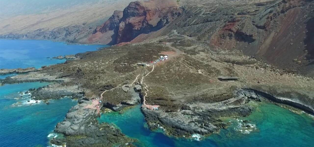 Dramatic volcanic coastline with turquoise water, rocky outcrops, and a winding mountain road leading to a white building on a barren hillside