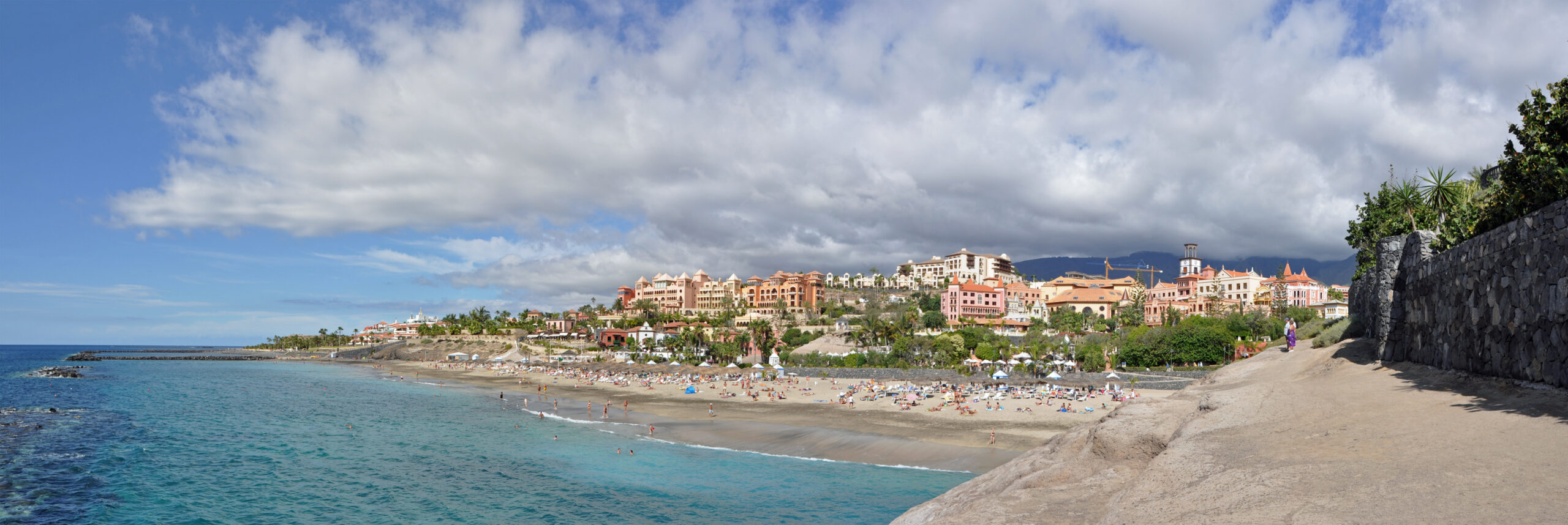 Sandy beach with turquoise water, sunbathers, and luxury resort buildings on a hillside under partly cloudy sky