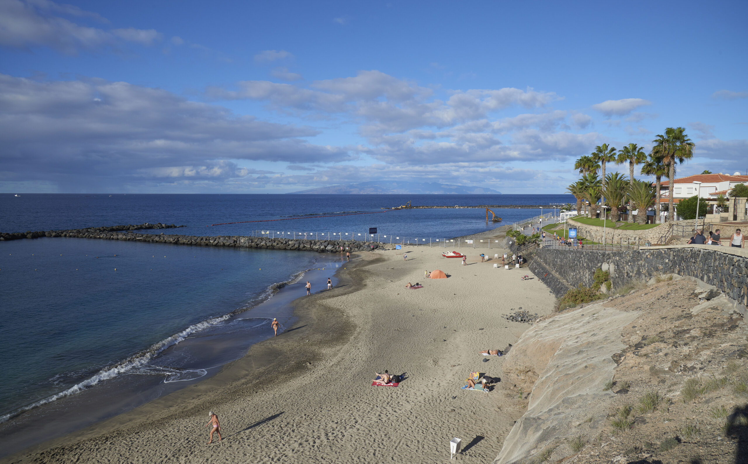 Sandy beach with scattered sunbathers, rocky breakwater, palm trees, and waterfront buildings under blue sky with white clouds
