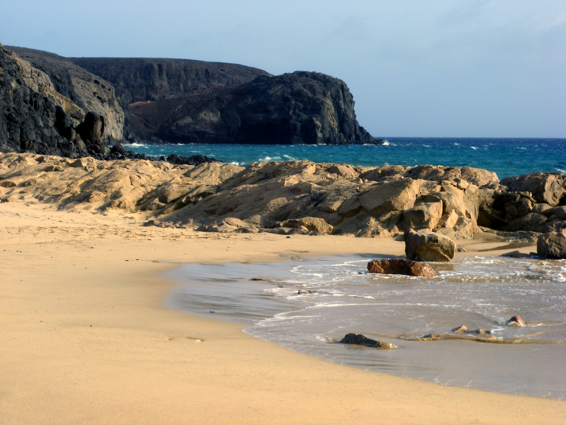 Playa del Pozo features golden sand, calm turquoise waters, and striking volcanic rock formations rising from the shoreline.