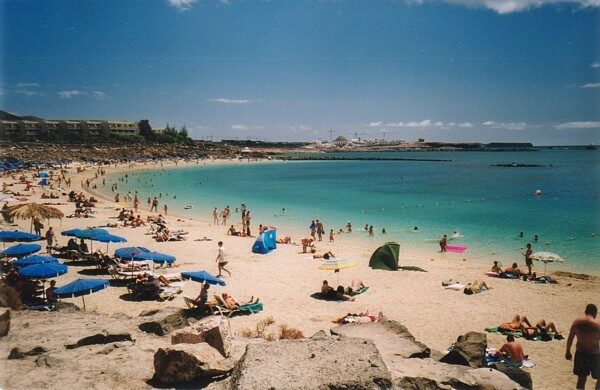 Wide sandy beach with turquoise water, blue umbrellas, sunbathers, and swimmers under clear blue sky with white clouds