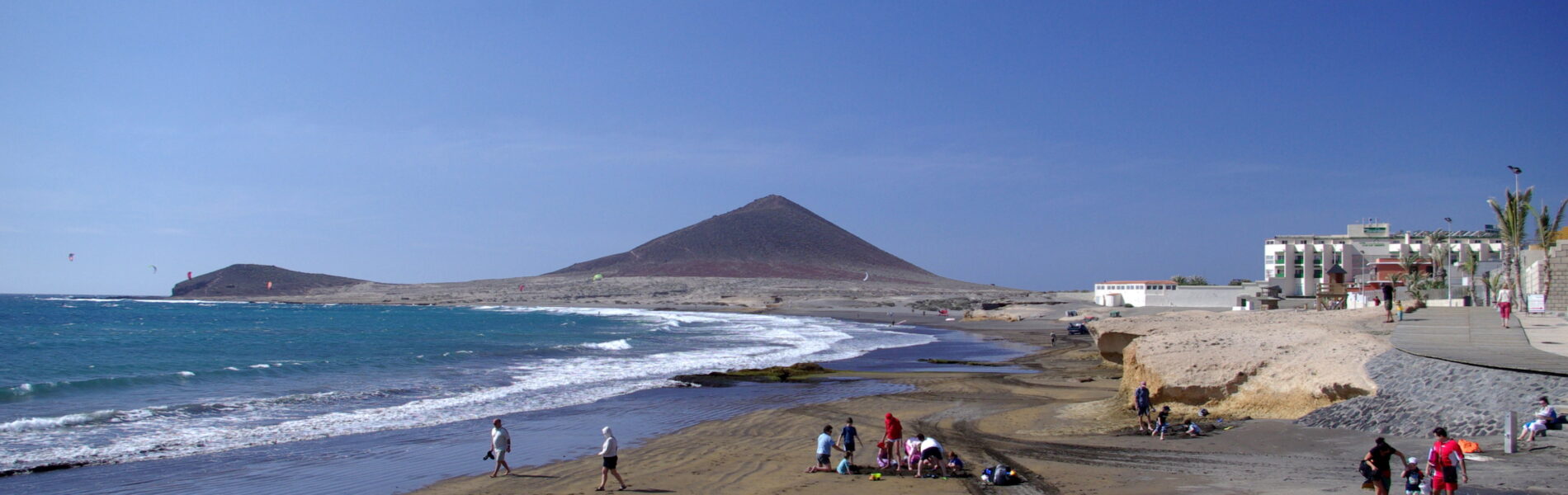 Sandy beach with waves and swimmers, volcanic cone mountain in distance, waterfront buildings and palm trees on right