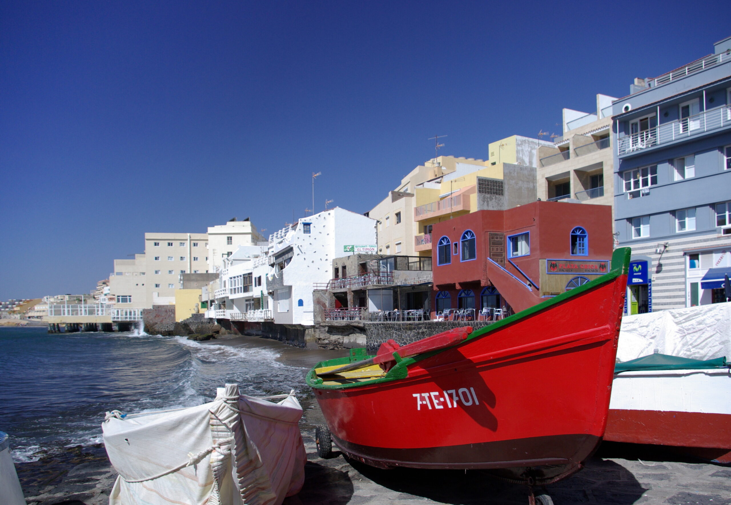 Red and green fishing boats on a beach with colorful waterfront buildings and clear blue sky