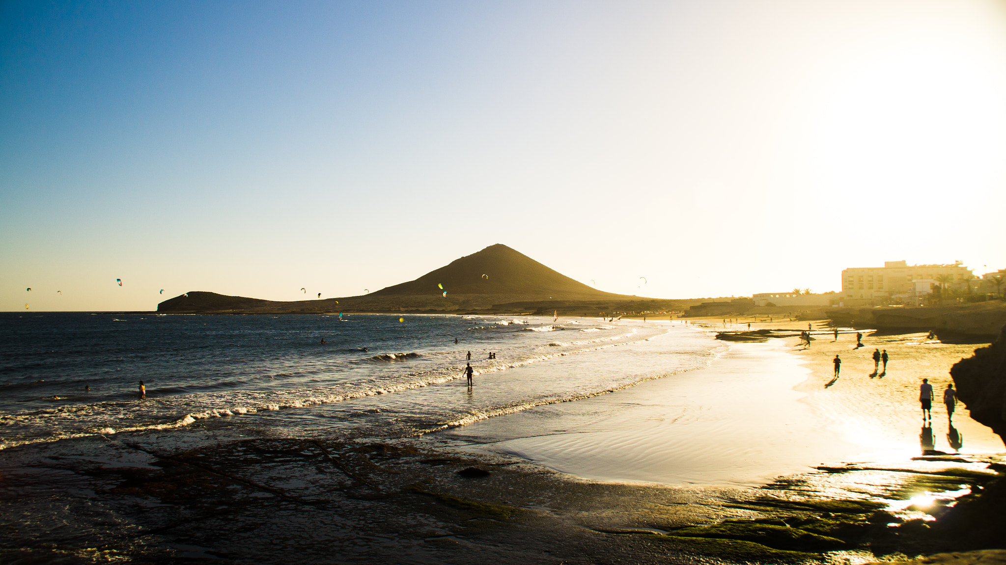 Sandy beach with shallow turquoise water and golden sunset light, a distinctive cone-shaped volcanic mountain across the bay, beachgoers swimming and wading, modern buildings on the right shoreline, kiteboarders on the water.
