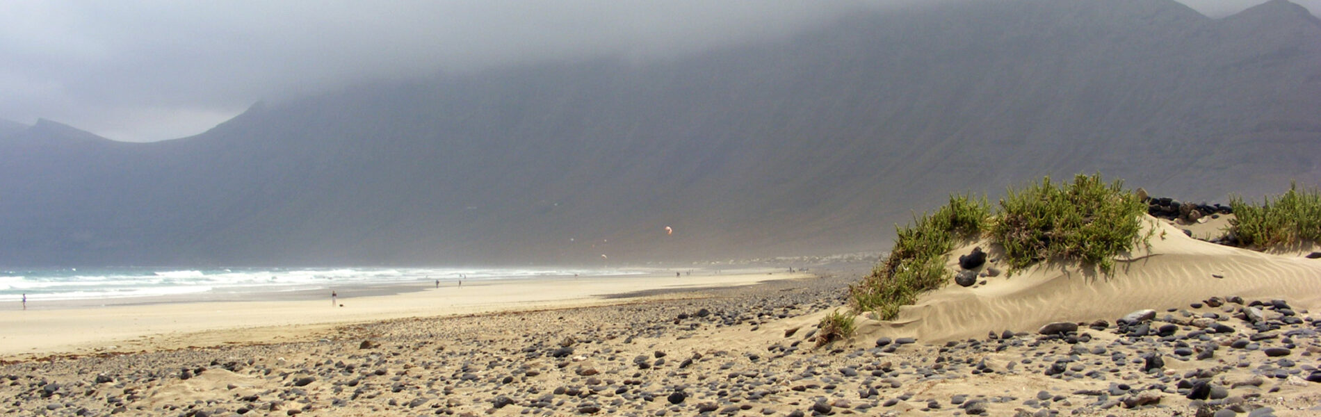 Sandy beach with dark volcanic rocks in foreground, gentle waves, sparse swimmers, and misty mountains rising behind under overcast sky.