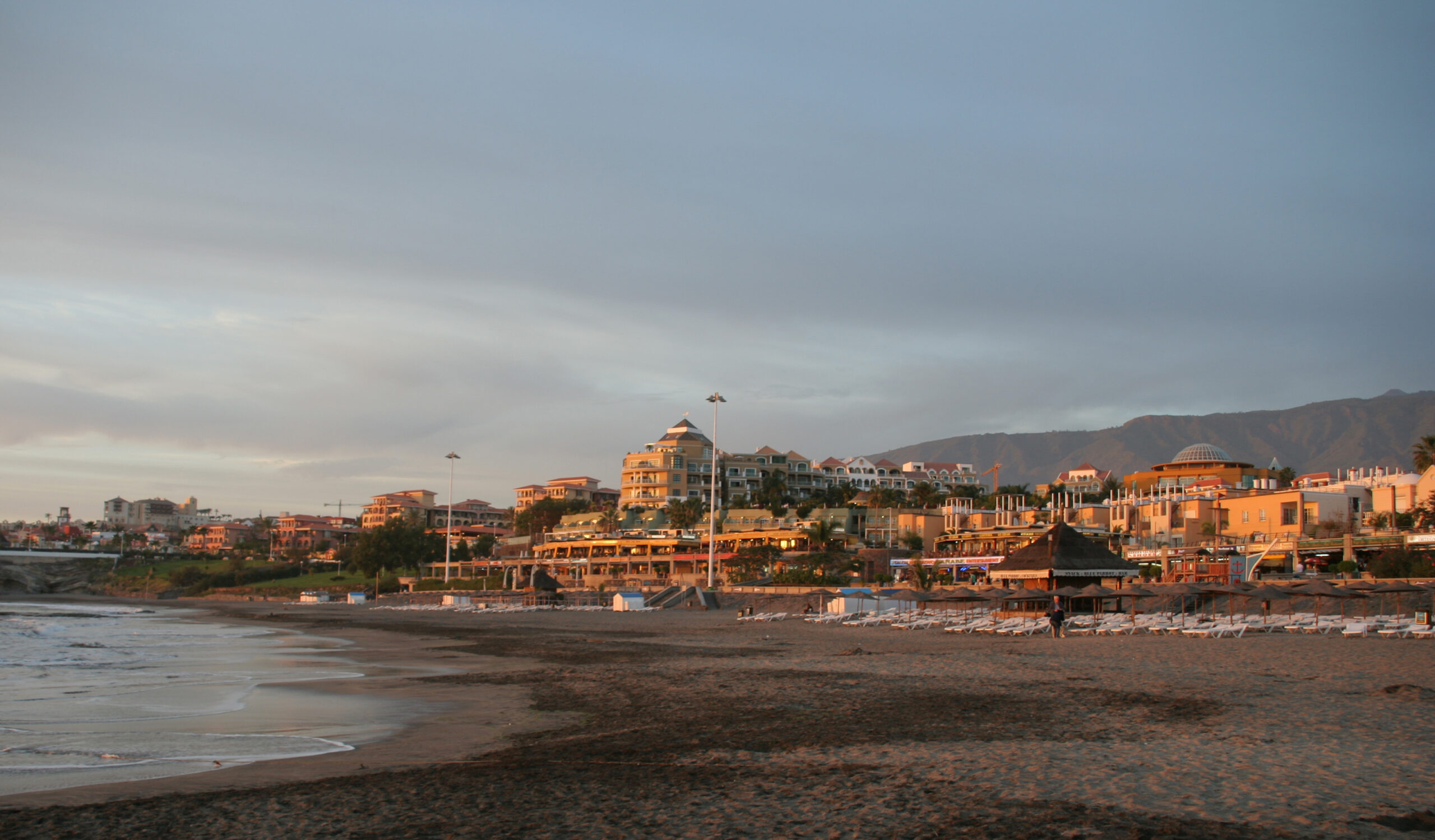 Beachfront resort buildings in golden light at sunset, with dark sand beach and gentle waves, mountains in background.