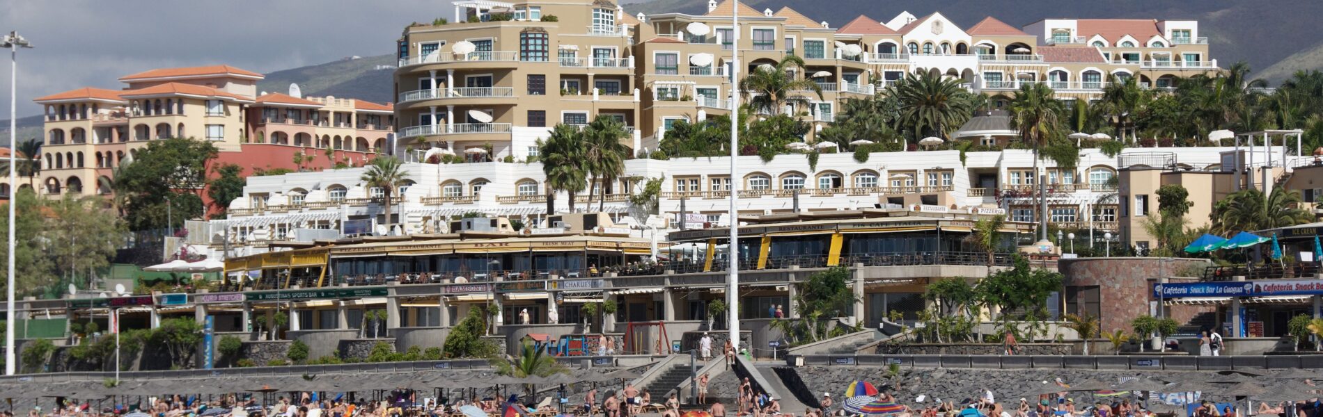 Crowded black sand beach with swimmers and sunbathers, backed by multi-story hotels and apartment buildings with palm trees, under partly cloudy sky and mountains.