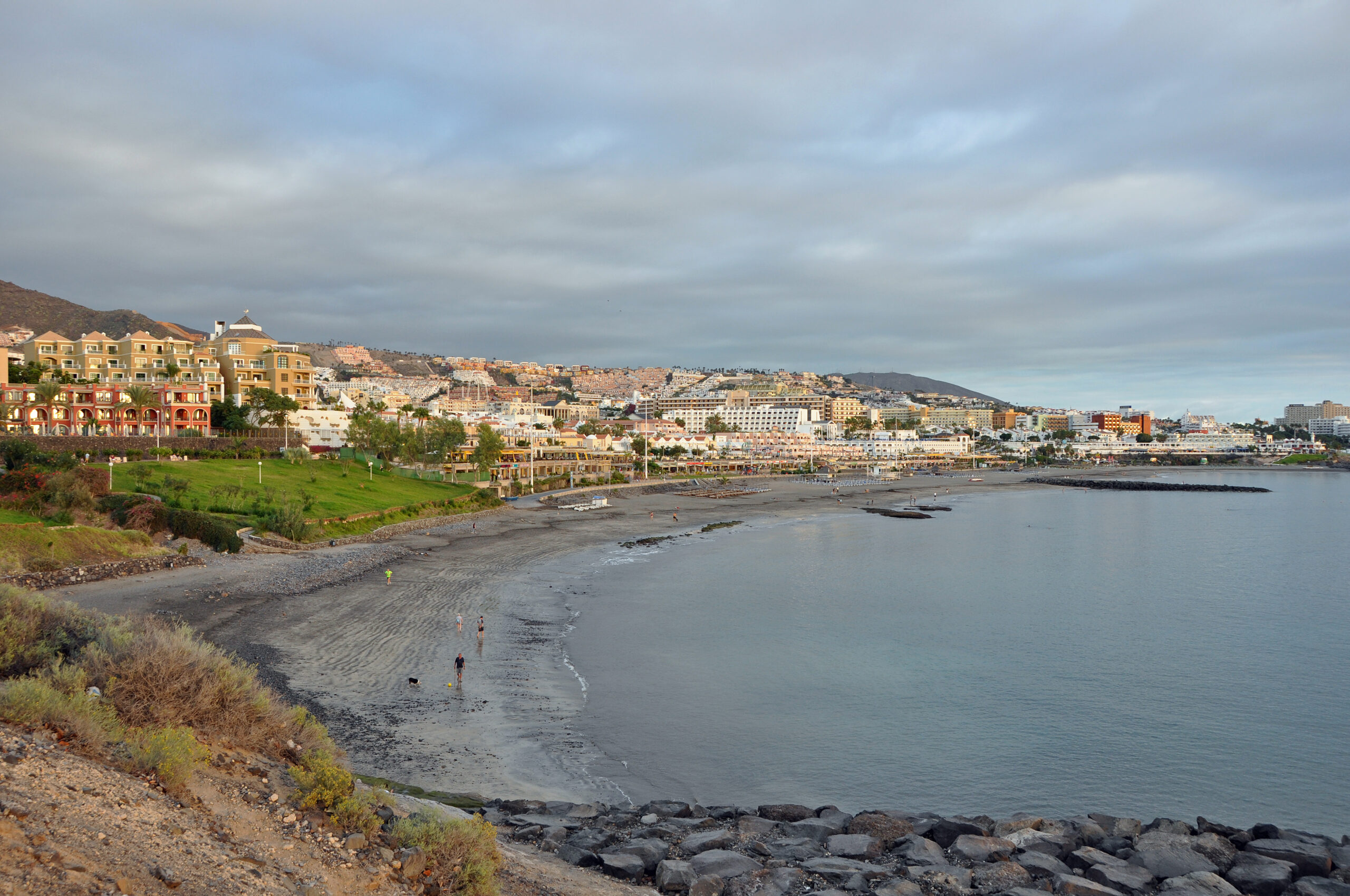 Curved sandy beach with dark volcanic sand bordered by green grass and colorful apartment buildings climbing a hillside under overcast sky