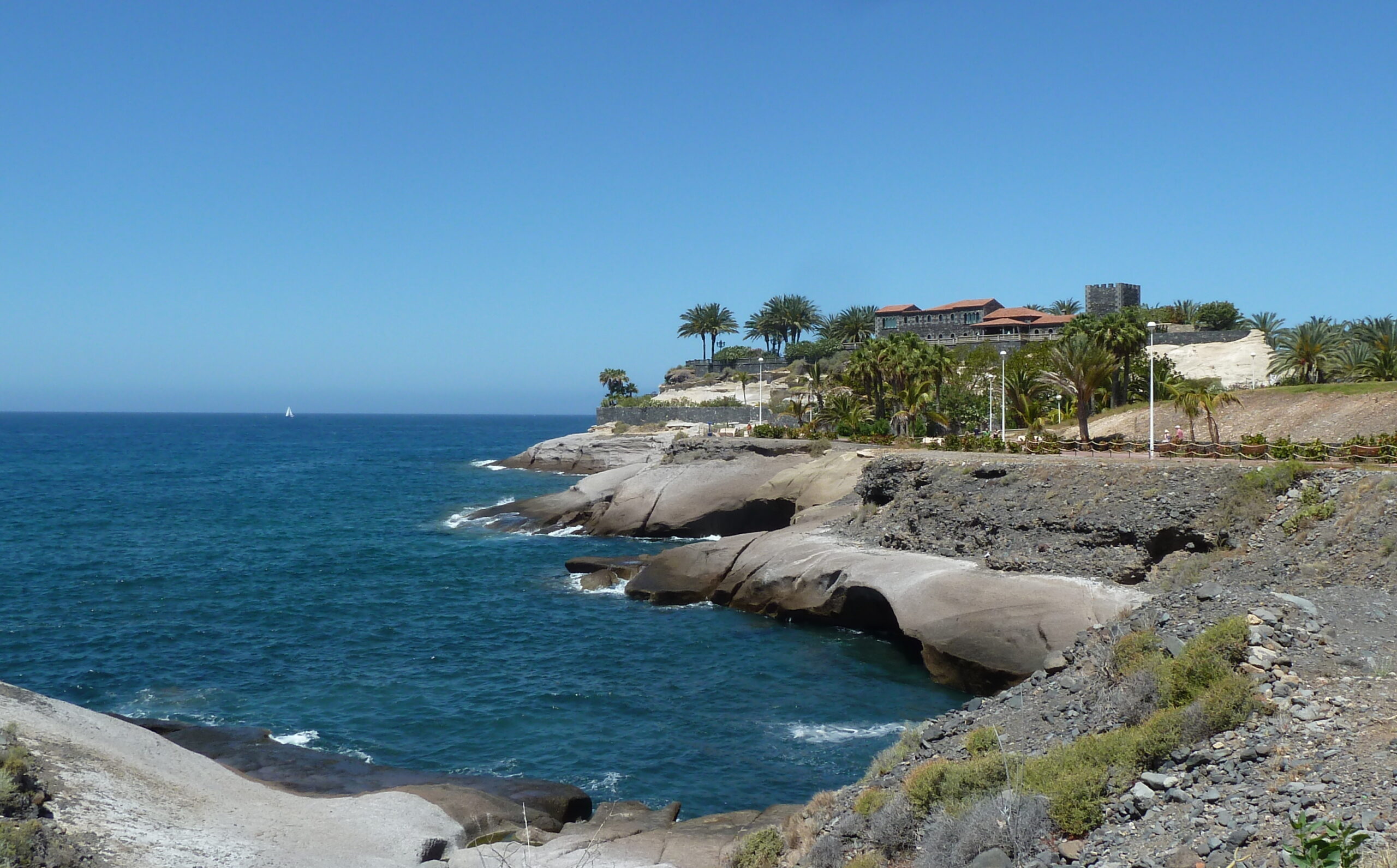 Coastal landscape with distinctive rounded granite boulders in turquoise water, palm trees, and a hilltop resort building under clear blue sky
