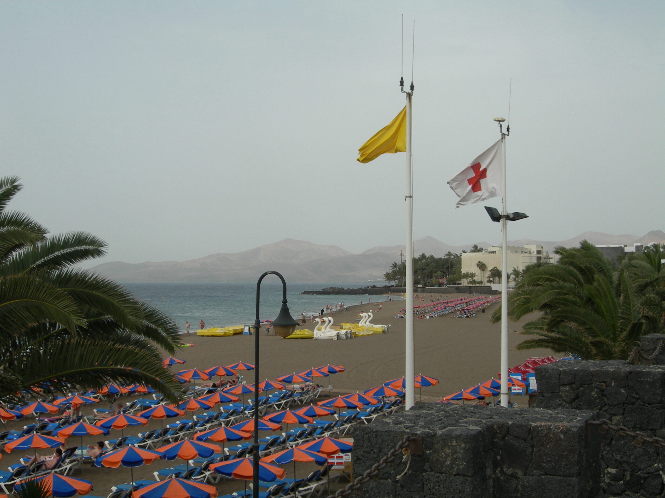Playa Grande in Puerto del Carmen features dark volcanic sand, colorful beach umbrellas, and views across the water toward Lanzarote's mountainous interior.