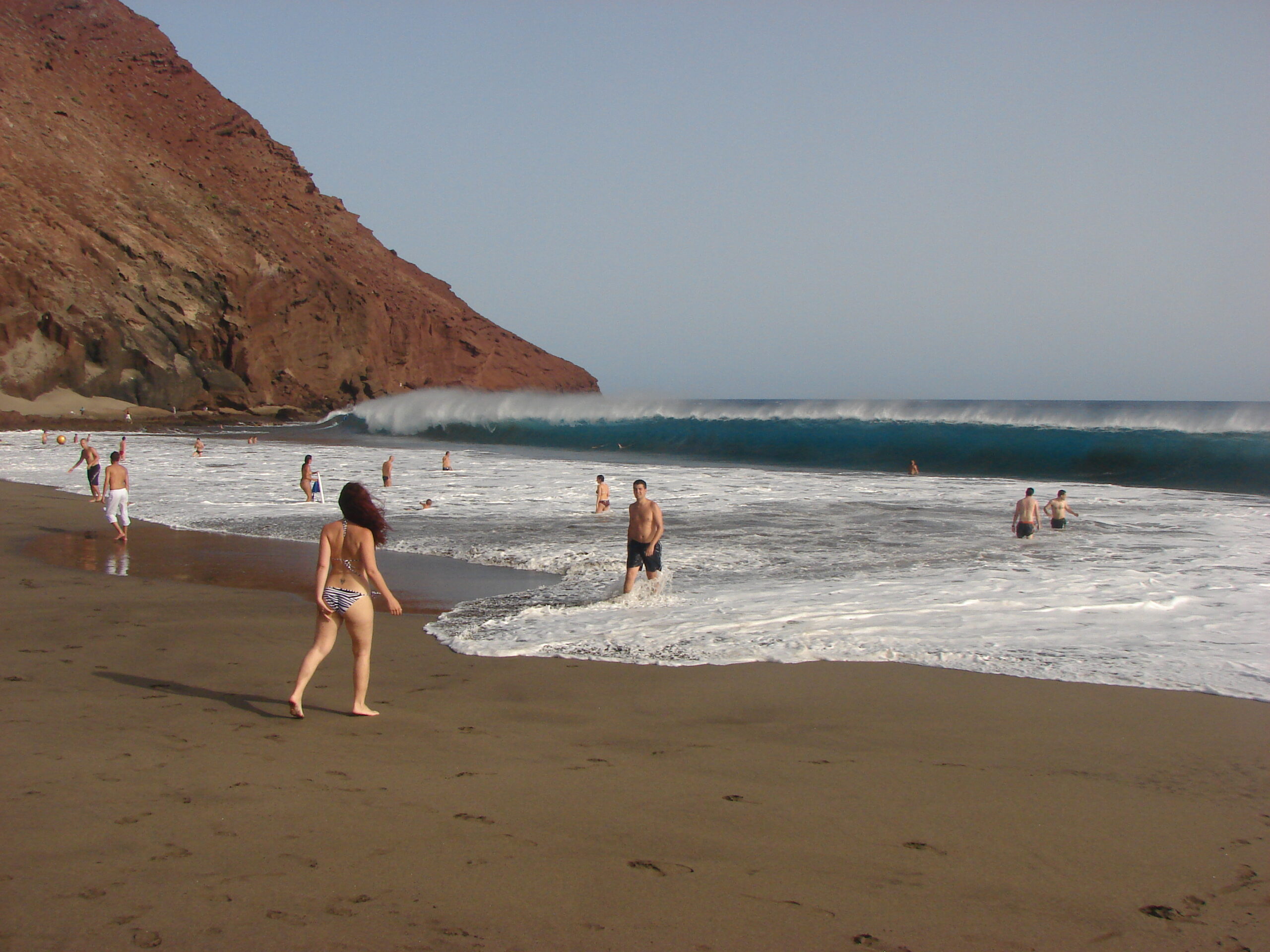 Swimmers and waders on a sandy beach with large waves rolling in, backed by a distinctive red volcanic cliff