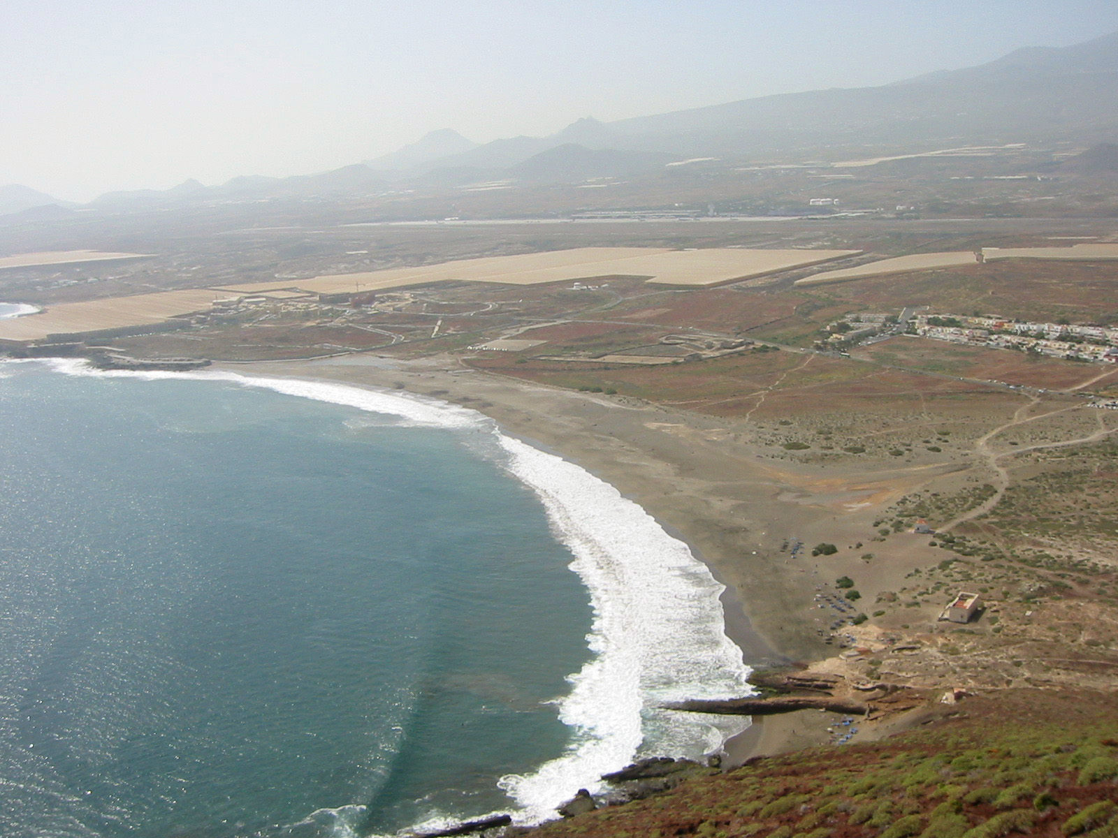Aerial view of a sandy beach with white-crested waves, volcanic coastline, and agricultural fields inland beneath misty mountains.
