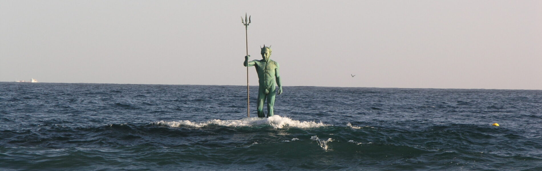 Green bronze statue of Neptune holding a trident, standing in shallow ocean waves at a beach.
