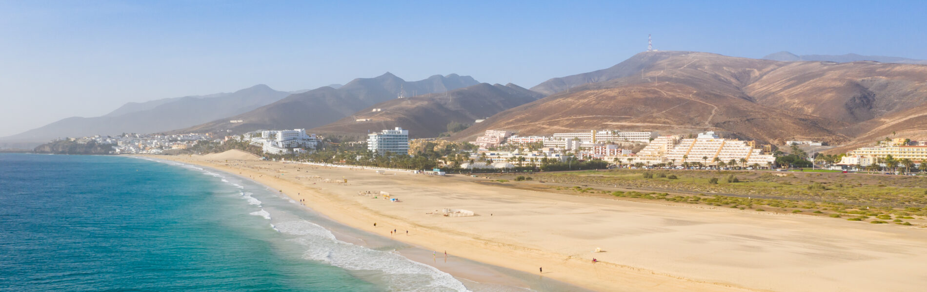 Wide sandy beach with turquoise water and white-capped waves, backed by low-rise resort hotels and arid mountains under clear blue sky