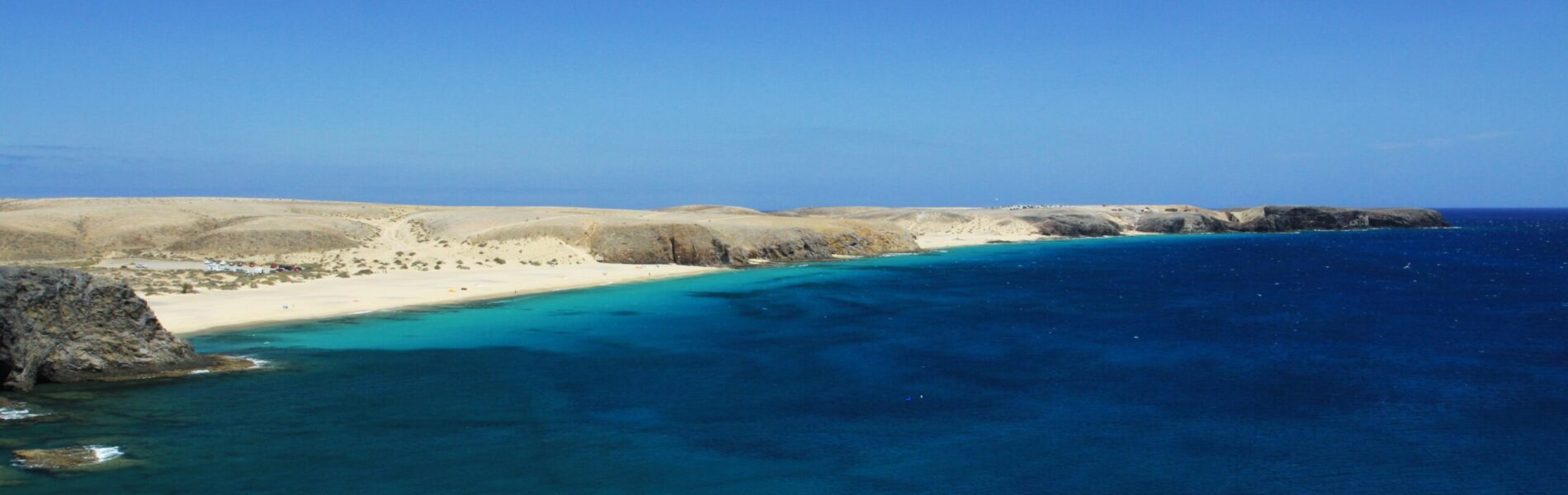 Turquoise water and golden sandy beach backed by arid hills under clear blue sky, viewed from above