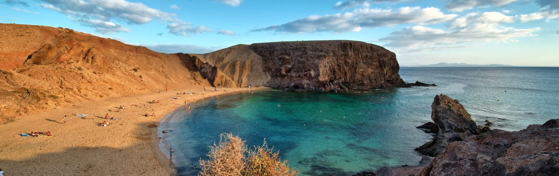 Secluded sandy beach with turquoise water framed by golden cliffs and dark volcanic rock formations under blue sky