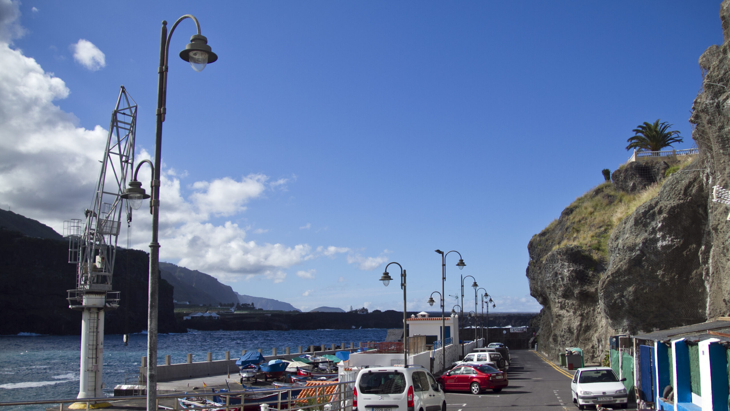 Coastal harbor with parked cars, vintage lamp posts, rocky cliffs, moored boats, and blue water under clear sky