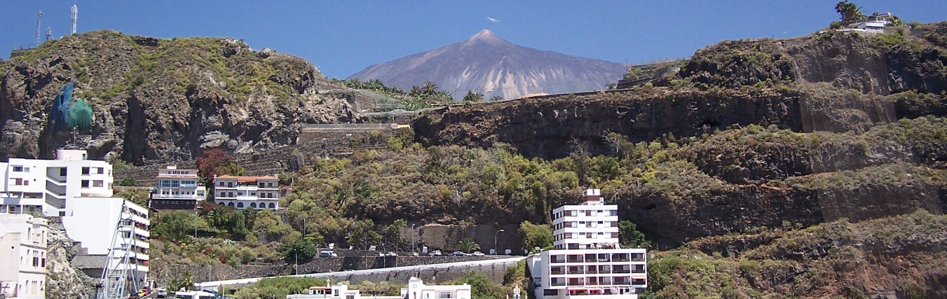 Coastal town nestled below volcanic cliffs with a cone-shaped volcano visible in the distance under clear blue sky
