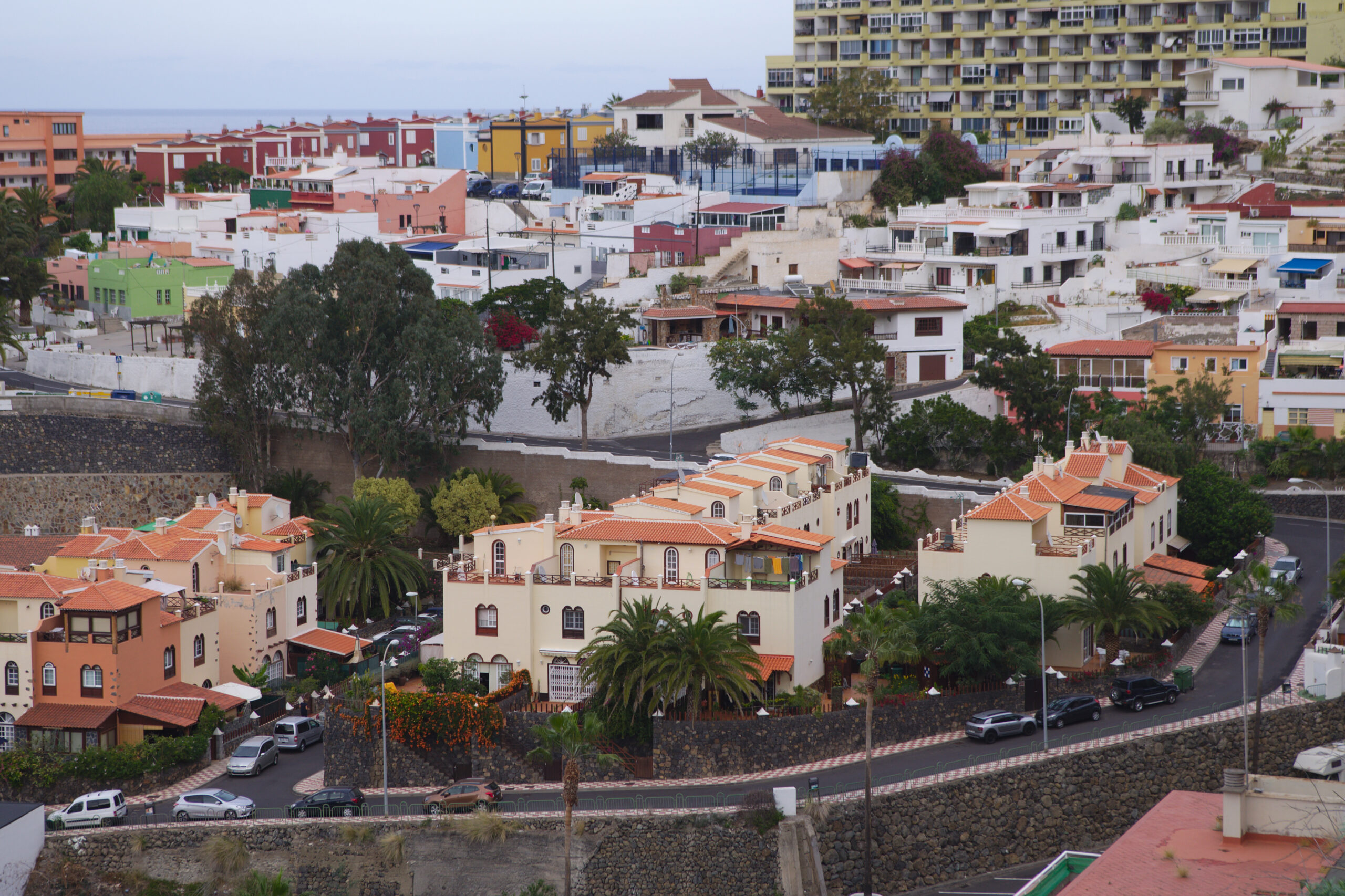 Colorful buildings with terracotta roofs cluster around a cream and orange colonial-style structure in a hillside coastal town, with palm trees and a stone wall visible.