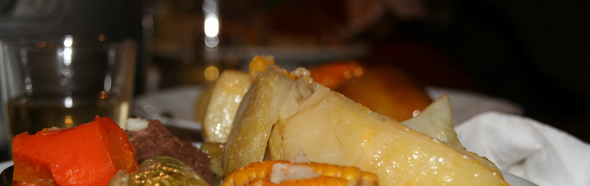 White plate of puchero canario with boiled potatoes, chickpeas, red pepper, and meat broth, served at a restaurant table with glasses of water and a blurred diner in background