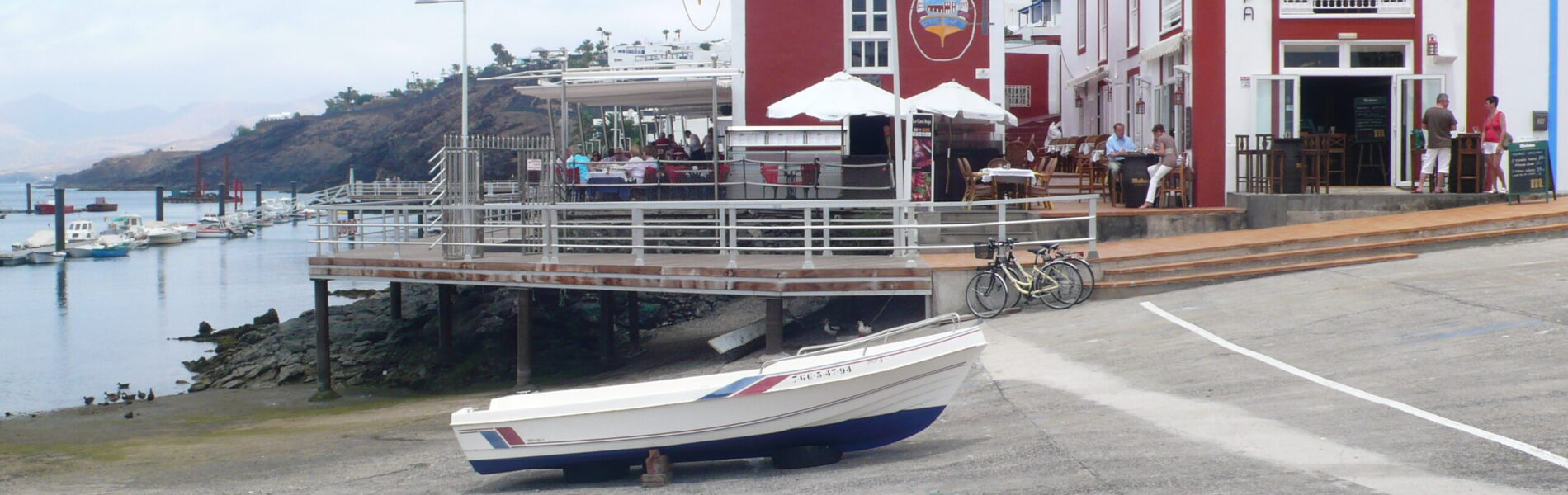 Waterfront promenade with red and white buildings, wooden boardwalk, moored fishing boat, and hillside in background under clear sky