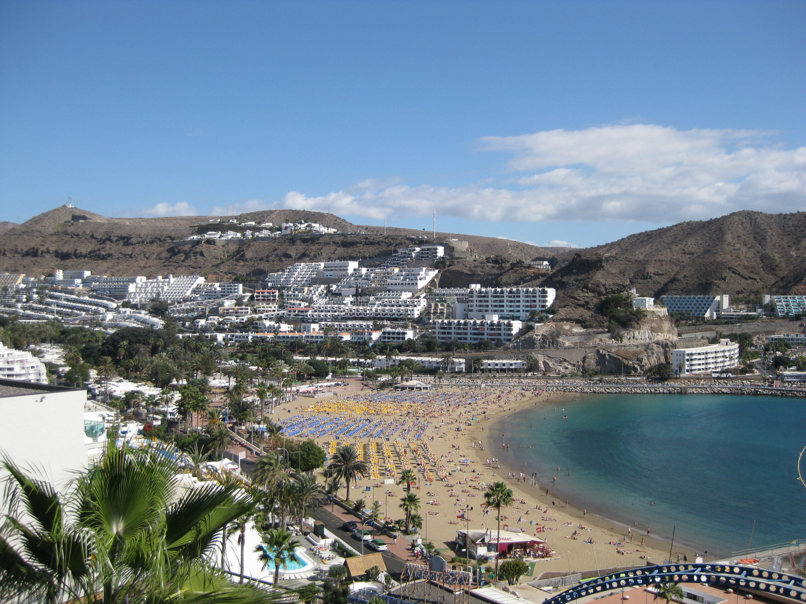 Puerto Rico Beach features a crescent of golden sand, rows of yellow beach umbrellas, and cascading white resort buildings terraced into the volcanic hills.