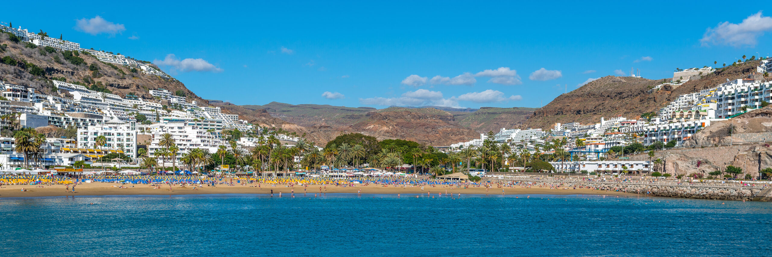 Puerto Rico beach stretches along the southern coast of Gran Canaria, framed by terraced white apartments and backed by dry, rocky hills.