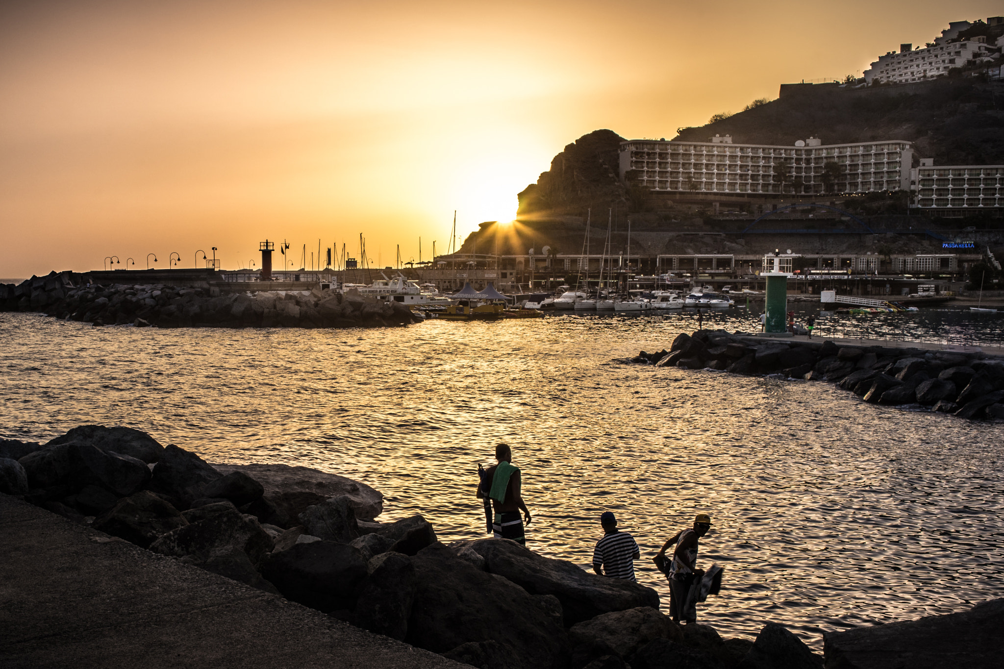 Puerto Rico Beach glows in golden sunset light, with swimmers enjoying the sheltered cove while the marina and clifftop hotels form the backdrop.